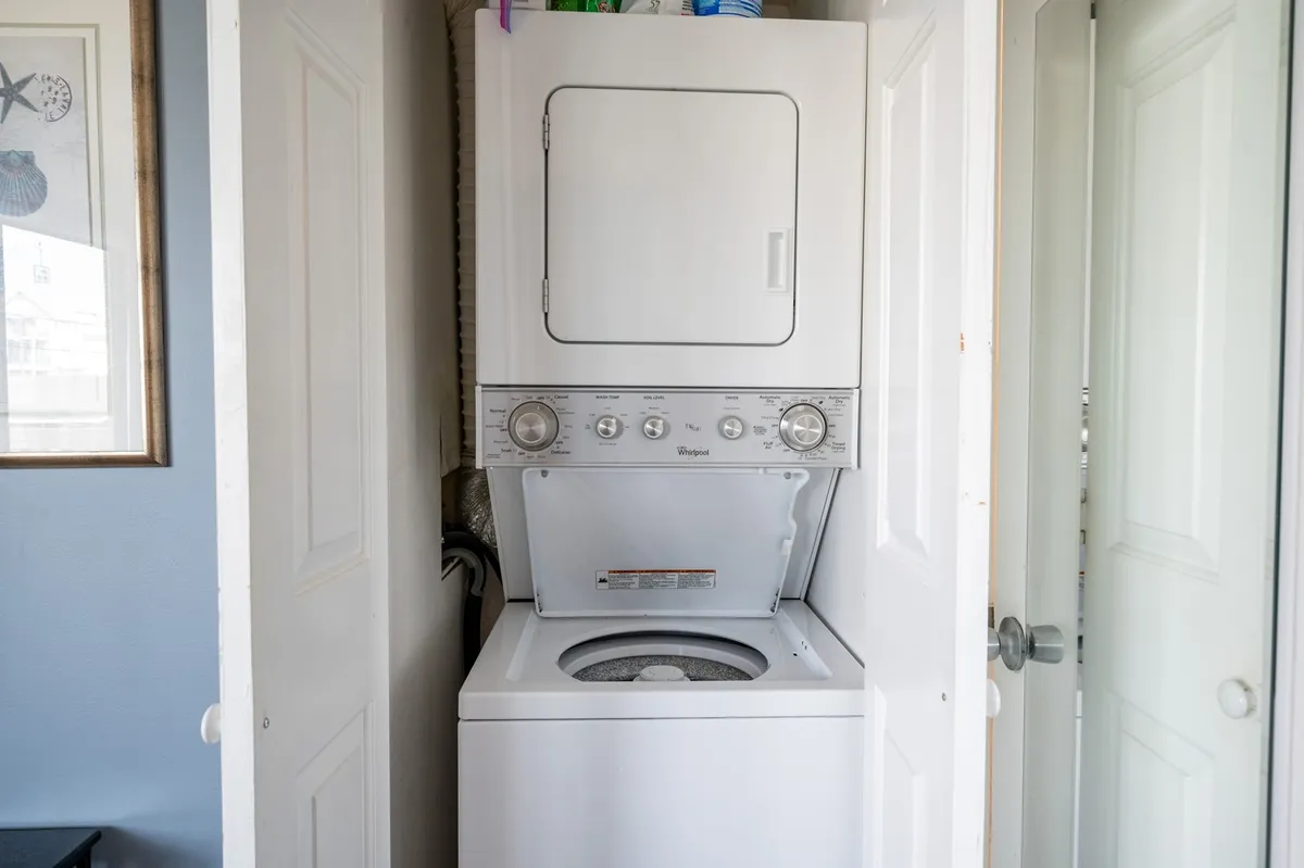 A practical laundry nook featuring a stacked washer and dryer, complemented by framed artwork and open doors. The space is designed for convenience and efficiency, making laundry tasks easy after days at the beach or pool.