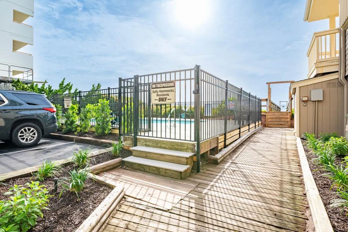 The image captures a sparkling community swimming pool surrounded by landscaped gardens, wooden steps leading into the area, and a fence with signage. Several lounge chairs are arranged for sunbathing and relaxation, with the pool inviting for a swim. The setting appears friendly and well-kept, ideal for guests to unwind or gather with friends.