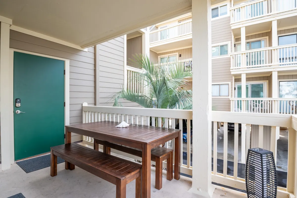 An inviting outdoor deck scene featuring a wooden table, green door leading inside, a palm plant, and comfortable outdoor seating. The setting is perfect for alfresco dining, morning coffee, or relaxed social gatherings.
