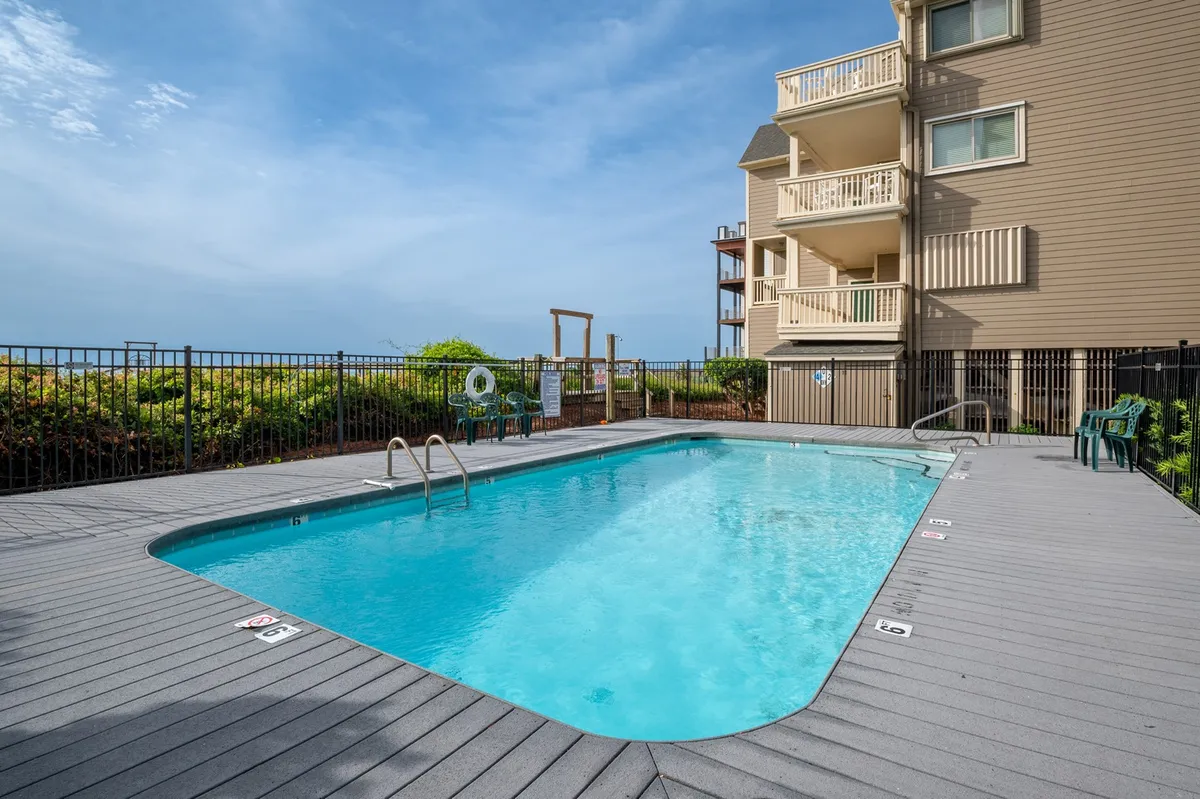 The image showcases a sparkling swimming pool surrounded by lush greenery and sun-soaked lounge chairs, with the condo's balcony overlooking the inviting pool area. The scene radiates tranquility and offers the perfect setting for sunset swims, relaxing poolside, and socializing with friends or family.
