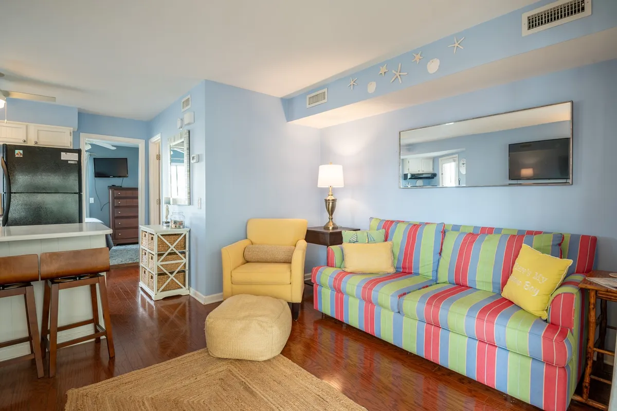 A bright, open interior with a striped couch, vibrant yellow armchair, decorative mirror, and a kitchen area in the background. The space exudes a playful, relaxed vibe with tropical colors and ample natural light streaming through large windows.