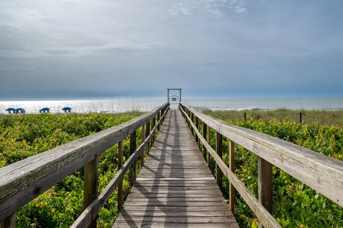 A wooden boardwalk leads over the dunes directly to the beach, flanked by blue beach chairs and an open view of the ocean. The peaceful setting emphasizes easy, private access to the shoreline and the tranquil atmosphere awaiting guests.