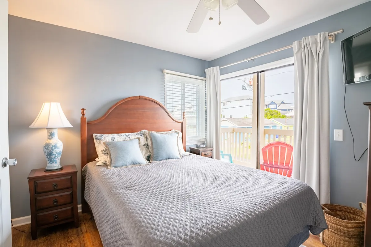 This image features a comfortable bed positioned beside a window that lets in ample natural light, with a lamp on the bedside and a glimpse of colorful chairs on an outdoor space just outside. The setting is simple yet inviting, evoking a sense of relaxation and personal comfort.