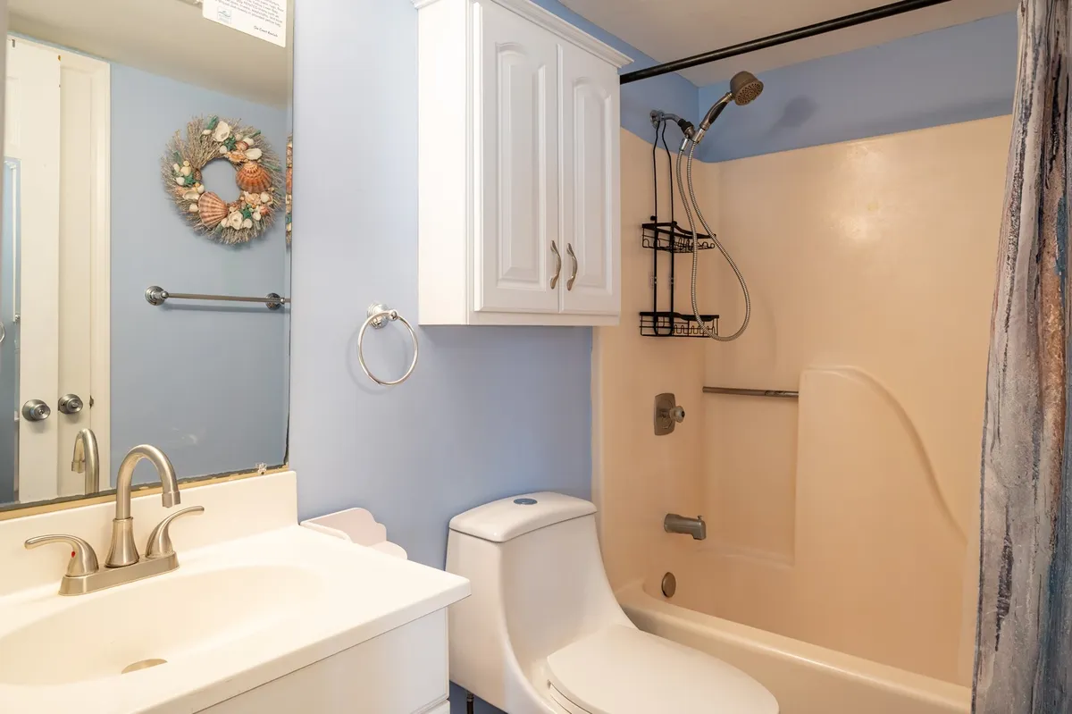A bright and serene ensuite bathroom is shown, featuring a clean sink area with a mirror, a neatly decorated wreath, a toilet, and a spacious shower area. The bathroom feels inviting and thoughtfully designed for relaxation and convenience.