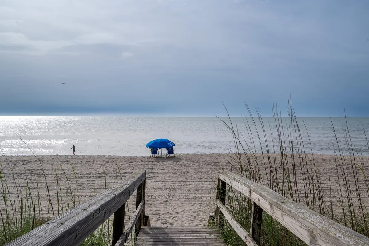 A sunlit wooden walkway leads over the sand dunes directly to a wide, inviting beach. A blue beach umbrella stands out against the soft sand, with a distant figure strolling by the ocean waves. Palm trees and the open sky create a romantic, relaxed seaside atmosphere perfect for couples.