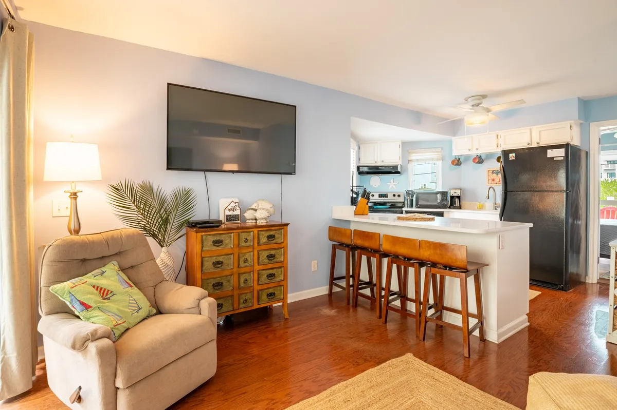 A welcoming living room scene featuring a comfortable armchair positioned near a TV, a kitchen island in the background, and a decorative cabinet. The space is bathed in natural light, with colorful, relaxed décor that hints at a tropical, laid-back atmosphere.