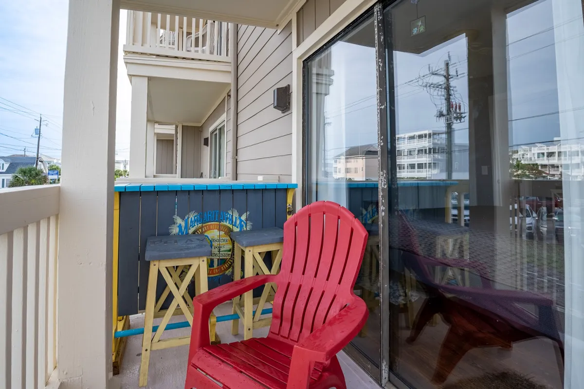 A cozy balcony with a vibrant red Adirondack chair and colorful railings overlooks a tranquil outdoor setting. A small wooden table invites you to enjoy a morning coffee or evening drink, with the soft glow of sunrise or sunset in the background. The scene feels intimate and inviting—ideal for couples seeking relaxation.